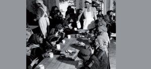 Et foto av samiske barn kledd i kofte spiser ved en langbord, mens norske kvinner med hvite forklær serverer mat